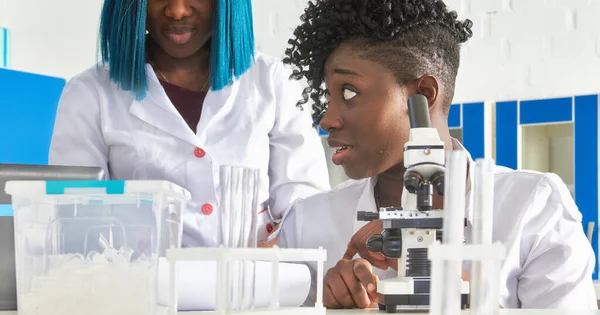 Genç ve çekici Afrikalı kadın bilim adamı, mezun ya da tıp öğrencisi test laboratuarı ya da üniversite biyoloji pratik kursunda hasta biyopsisi örneklerini mikroskopla gözlemler. Meslektaşıyla konuşuyor..