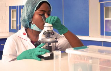 Afrikalı tıp öğrencisi, koruyucu beyaz önlüklü kadın bilim adamı plastik şapka, eldiven, maske laboratuvarda mikroskopla çalışıyor. Hasta biyopsi örneklerinin analizi. Anatomist çalışıyor