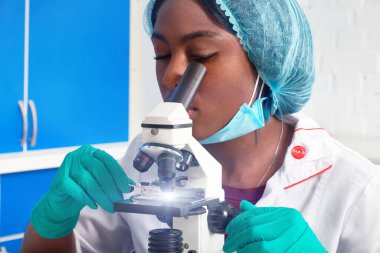 Koruyucu beyaz önlüklü, plastik şapkalı, eldivenli, maskeli genç Afrikalı kadın bilim adamı modern laboratuarda mikroskopla çalışıyor. Hasta biyopsi örneklerini analiz et, kanser teşhisi koy.