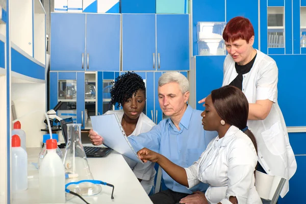 病院の検査室で科学者や医療の多文化チームは Covid 19コロナウイルス検査のためのテストシステムを議論します ヨーロッパのシニア同僚やジュニアアフリカの研究室メンバーは 一緒に動作します — ストック写真
