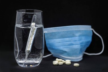 Bir bardak su, termometre, tıbbi maske ve siyah arka planda izole edilmiş haplar..
