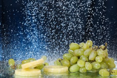 Suyun altında koyu mavi arkaplanda yeşil üzüm damlaları var. Sağlıklı bir yaşam tarzı. Multivitamin kokteyli. Yaz diyeti. Suyun altında meyve seti. Düşen üzüm ve ananas dilimleri