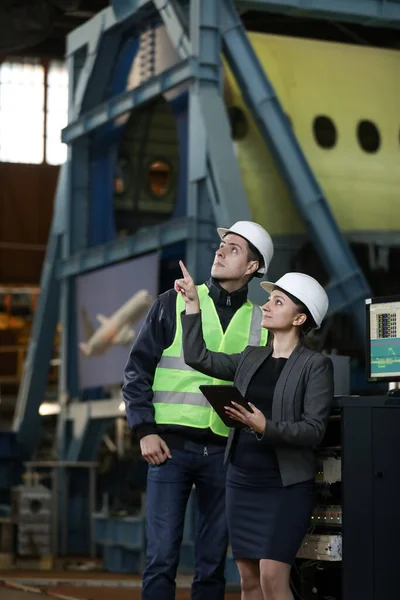 Portrait of a female factory manager in a white hard hat and business suit and factory engineer in work clothes. Controlling the work process at the airplane manufacturer.