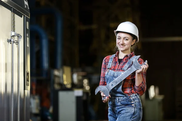 Young girl in a work dress and white hard hat holding big wrench and hummer in a factory. Woman in a work uniform. Working process.