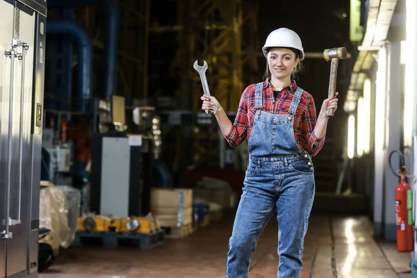 Young girl in a work dress and white hard hat holding big wrench and hummer in a factory. Woman in a work uniform. Working process.