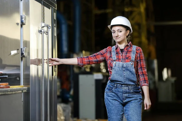 Young girl in a work dress and white hard hat in a factory. Woman in a work uniform. Working process.