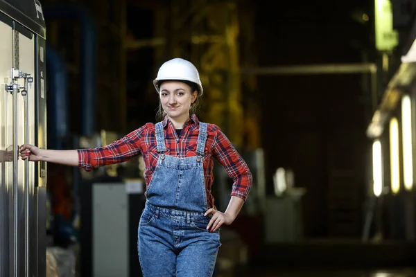 Young girl in a work dress and white hard hat in a factory. Woman in a work uniform. Working process.