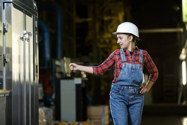 Young girl in a work dress and white hard hat in a factory. Woman in a work uniform. Working process. Girl open the refrigerator compartment.