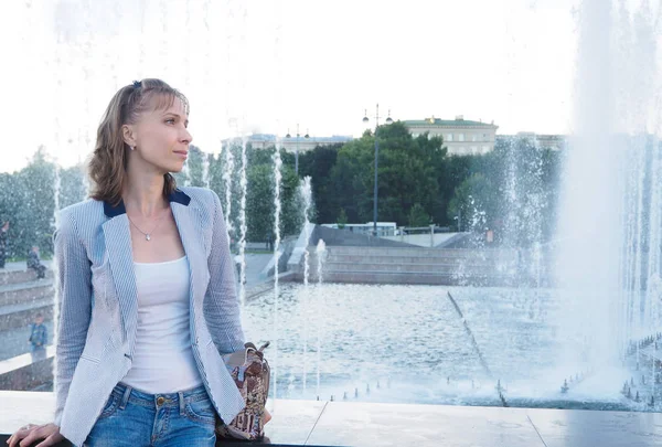 Uma mulher com um casaco leve que descansa ao pé da fonte. Mulher feliz livre desfrutando fonte da cidade no dia de verão . — Fotografia de Stock