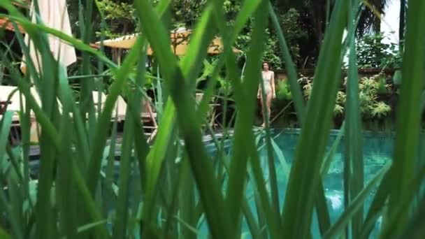 Una vista attraverso le foglie: una ragazza cammina a bordo piscina ai tropici — Video Stock