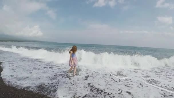 La ola se rompe en la orilla y grandes aerosoles vuelan sobre la chica en la playa de arena negra — Vídeos de Stock