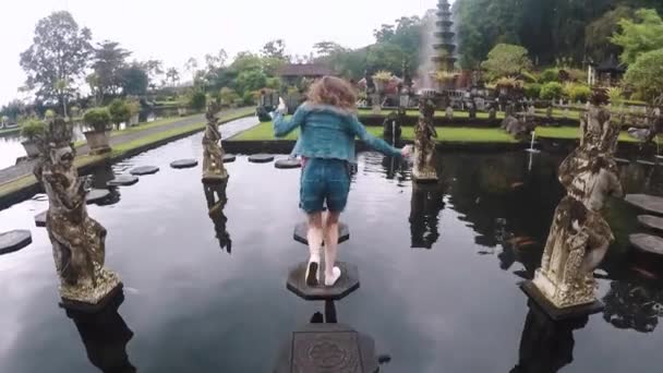 Камера идет вверх: молодая женщина бежит вдоль плитки в пруду в красивом балийском храме Tirta Gangga на Бали, замедленная съемка — стоковое видео