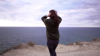 Güzel bir genç bayan sporcu rüzgarlı bir günde uçurumun kenarına yürür. Spor eşofman üstü, sert rüzgar bulaşık makineleri kızlar uzun saçlar.