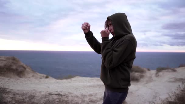 La práctica de la autodefensa. Entrenamiento de boxeo de mujer joven fitness al atardecer en un acantilado. Mujer deportiva independiente en una sudadera y polainas — Vídeos de Stock