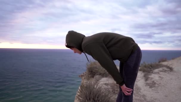 Een vrouw voelt zich moe na een zware training buiten op een klif. Fitness, moeilijke oefeningen. Onderbreking van de training, het meisje herstelt de ademhaling — Stockvideo