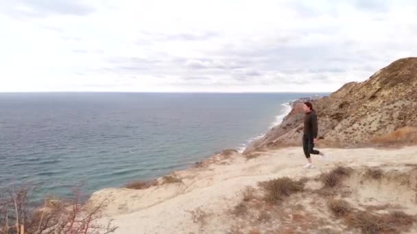 Luchtfoto - een jonge vrouw voelt vrijheid, staande op de rand van een klif boven de oceaan. Sportvrouw buiten in een hoody en losin — Stockvideo