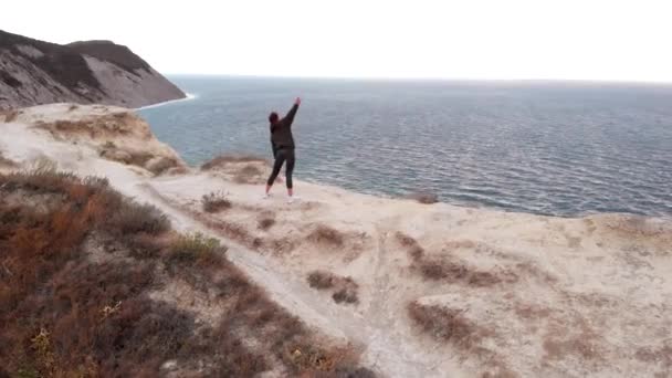 Εναέρια άποψη μιας γυναίκας που κάνει ασκήσεις τεντώματος στο φόντο του ωκεανού. Ευέλικτο κορίτσι κάνει υπαίθρια προπόνηση σε ένα βράχο — Αρχείο Βίντεο