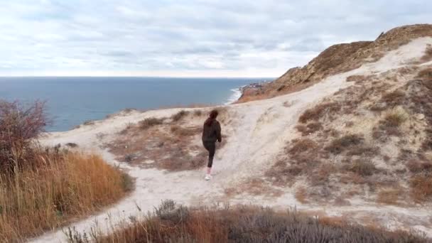 Z lotu ptaka widać trekking, kobieta jest zaangażowana we wspinaczkę na wzgórze z widokiem na morze. Aktywna kobieta w odzieży sportowej — Wideo stockowe