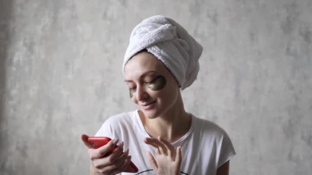 Eine glückliche Frau mit Pflaster unter den Augen blickt auf das Telefon und lacht. Feuchtigkeitsspendende und straffende geschwollene Haut — Stockvideo