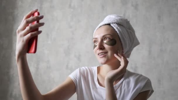 Meisje is thuis aan het vloggen. Een jonge vrouw met vlekken onder haar ogen en een handdoek kijkt naar de camera van een mobiele telefoon. Hydrateren van de huid en tillen — Stockvideo