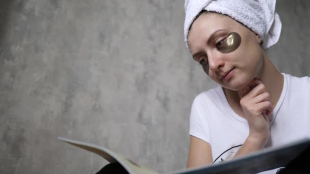 Cuidados com a pele, manchas sob os olhos de uma jovem mulher. Menina lendo uma revista em casa em uma toalha. Procedimento de cosmética de elevação e bem-estar — Vídeo de Stock