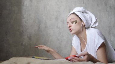 Cilt bakımı ve kulaklıkla müzik dinlemek. Akıllı telefonlu genç bir kadın sesi seviyor ve kanepede dergi okuyor.