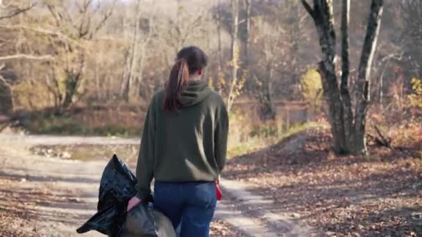 Egy női önkéntes szemeteszsákot és kesztyűt tart a kezében, miközben szemetet gyűjt. Tracking shot, őszi erdő, visszapillantás — Stock videók