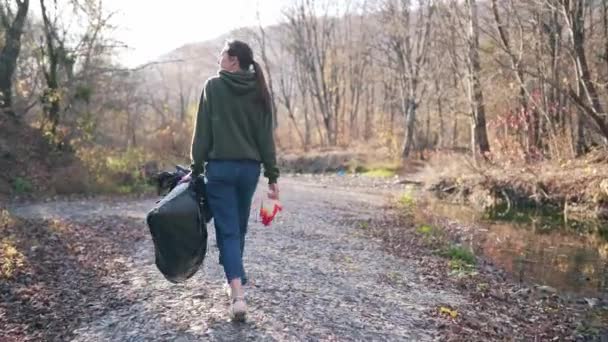 Περιβαλλοντική ρύπανση, εθελοντισμός έννοια. Μια νεαρή γυναίκα με μια μαύρη πλαστική σακούλα και γάντια στα χέρια της περπατά στη φύση κοντά στο ποτάμι — Αρχείο Βίντεο