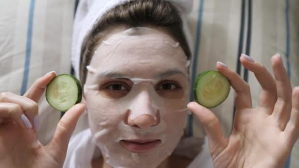 Μια νεαρή γυναίκα με μια μάσκα καθαρισμού και μια πετσέτα κοιτάζει την κάμερα και κρατά γύρω από αγγούρια στα χέρια της. Ευεξία, προϊόν ομορφιάς και περιποίηση δέρματος — Αρχείο Βίντεο