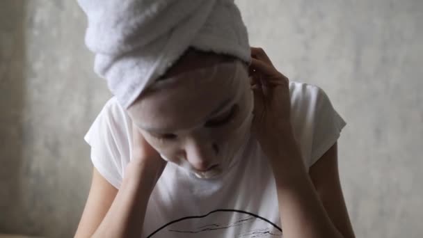 Blanke vrouw met een hydraterend masker op haar gezicht knoopt een witte handdoek op haar hoofd en kijkt naar de camera. Reiniging van het gezicht, verjonging, make-up en ontspannen — Stockvideo