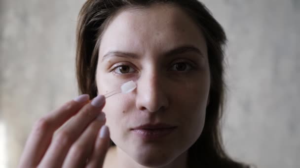 Bella giovane donna sta applicando una crema idratante trasparente contro le rughe sul suo viso. Cura del viso, pelle naturale, cosmetici — Video Stock