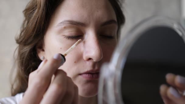 Una giovane donna applica una macchia di correttore con una spazzola sotto gli occhi e guarda in un piccolo specchio. Trucco di base, fondazione — Video Stock