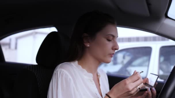 Maquillage dans la voiture. Portrait d'une jolie jeune femme en chemisier blanc soufflant sur un pinceau pour appliquer un fard à paupières — Video