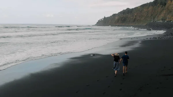 Krásný filmový záběr. Mladý pár běží za ruku podél černé sopečné písečné pláže. Letecký pohled na Tenerife, Španělsko — Stock fotografie