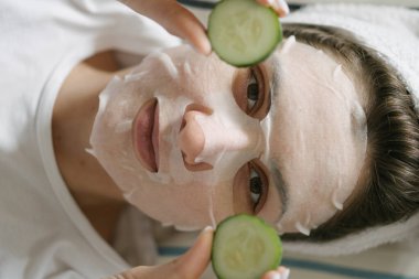 Temizleyici maske ve havlu takmış genç bir kadın kameraya bakar ve elinde yuvarlak salatalıklar tutar. Sağlık, güzellik ürünleri ve cilt bakımı