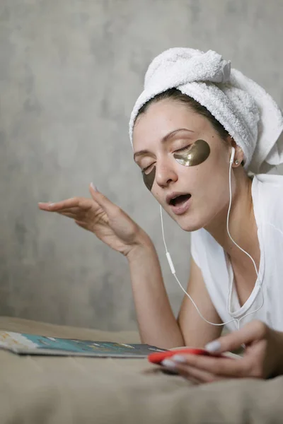 Atrakcyjna dziewczyna z ręcznikiem na głowie i łatami czyta magazyn i słucha muzyki. Kosmetyki nawilżające. Relaksujący styl życia — Zdjęcie stockowe