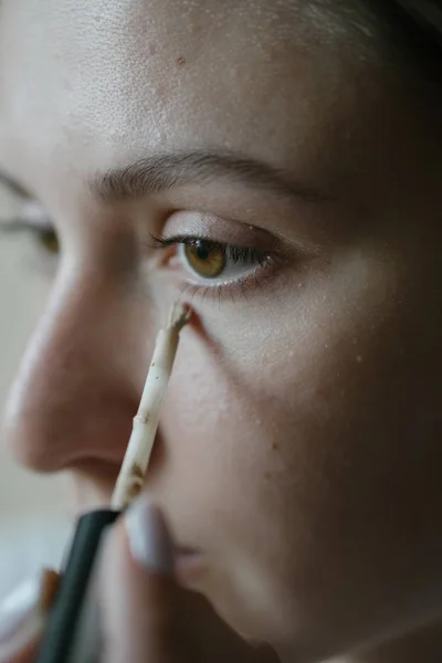 Μια νεαρή γυναίκα απλώνει ένα επίχρισμα κονσίλερ με μια βούρτσα κάτω από το μάτι της. Βασικό μακιγιάζ, θεμέλιο — Φωτογραφία Αρχείου