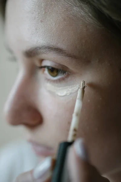 Toepassing van concealer onder het vrouwelijke oog close-up. Een blank meisje brengt een beige tinting foundation aan op de huid onder haar ogen. Vloeibare cosmetica — Stockfoto