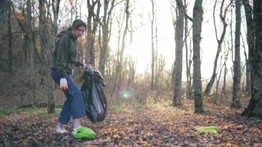 Güzel bir kız ağaçların ve güneş ışığının arka planına yaslanır ve plastiğin doğasını temizler. Temiz açık alan