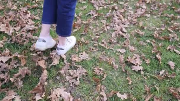 En tjej går på gräsmattan i jeans och gympaskor, varm vinter. Anklar utan strumpor i höstväder — Stockvideo