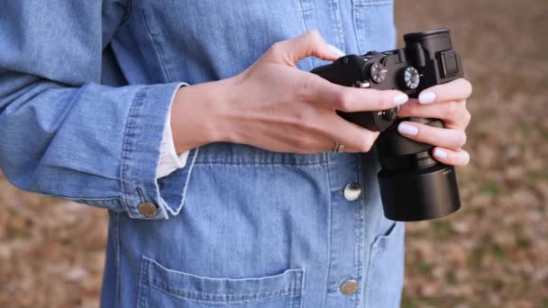 Schwarze Kamera in weiblichen Händen, Nahaufnahme. eine Frau in Jeanskleidung betrachtet und wählt das Filmmaterial auf einer spiegellosen Kamera auf der Straße — Stockvideo