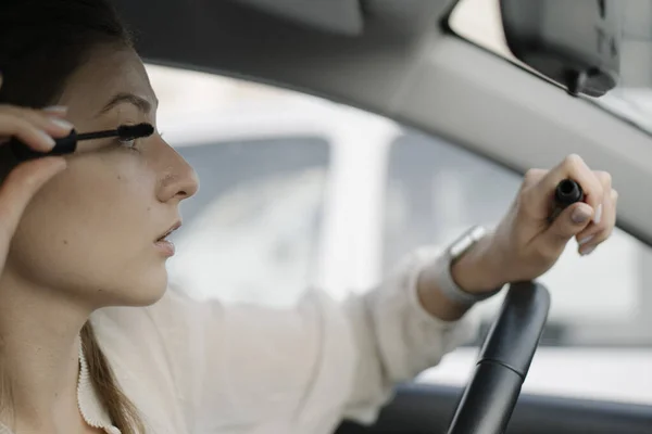 Eye brush, shadows, make-up in the car. A young woman looks in the mirror inside the car and applies make-up before a business meeting