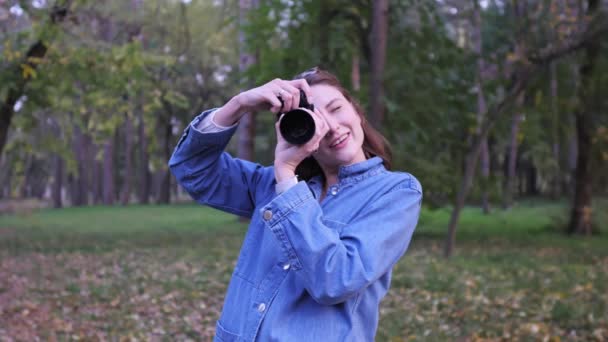 Menina fotógrafo hipster bonito tira uma foto em uma câmera profissional e sorri. Retrato no parque de outono, contra o fundo das árvores e folhas caídas — Vídeo de Stock
