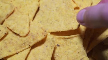 The hand takes one nachos, a macro static shot. The concept of unhealthy and fatty junk food - a mountain of yellow triangular chips close-up