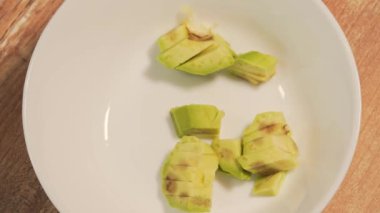 Kadınların elleri olgun avokado dilimlerini beyaz bir tabağa koyar. Vejetaryen salatası, yakın plan, üst manzara.