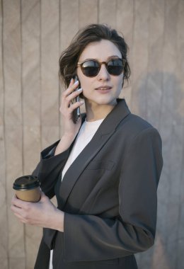 Şehir geçmişine karşı takım elbiseli güzel beyaz bir kadının portresi. Kız telefonda konuşuyor ve elinde bir fincan kahve tutuyor.