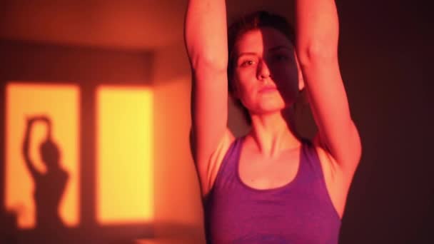 A beautiful girl does stretching exercises at sunset in the home room and looks into the camera. Sunset light, silhouette of a slender girl engaged in sports — Stock Video