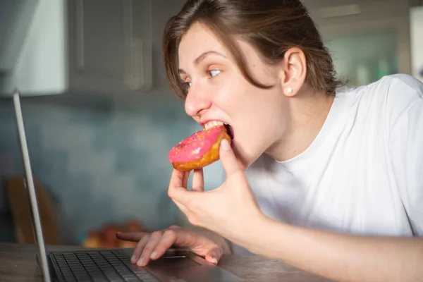 若い女性はピンクのドーナツを食べ、台所のラップトップを見ています。リモートワークと家庭教育をオンラインで。フリーランスの女の子は期限中にジャンクフードを食べる — ストック写真