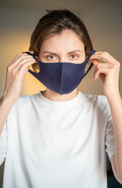 Genç bir kadın evde tekrar kullanılabilir koruyucu bir maske takıyor ve kameraya bakıyor. Coronavirus ve enfeksiyondan korunma. Covid-19 sırasında dışarı çıkmadan önce maskeli bir kızın portresi.
