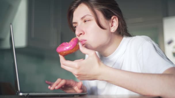 Uma jovem come um donut rosa e olha para um laptop em sua cozinha. Trabalho remoto e educação em casa online. Uma menina freelance comer junk food durante o prazo — Vídeo de Stock
